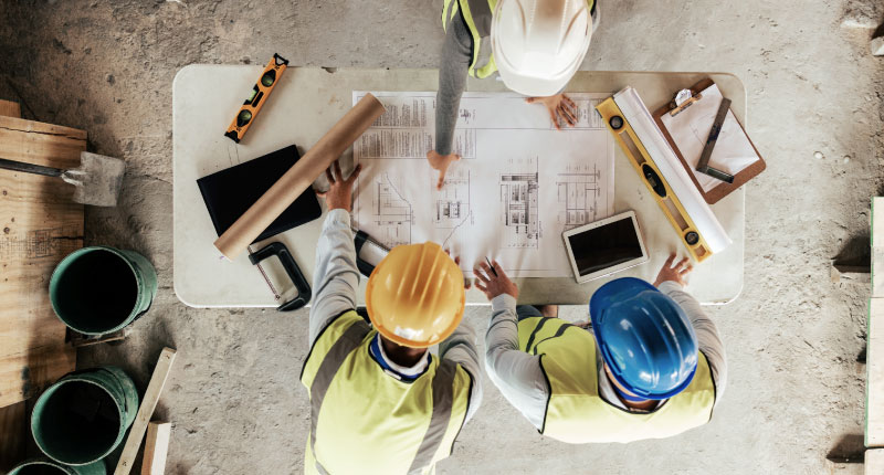 Top view of three construction workers looking at a blueprint kept on a table with other tools